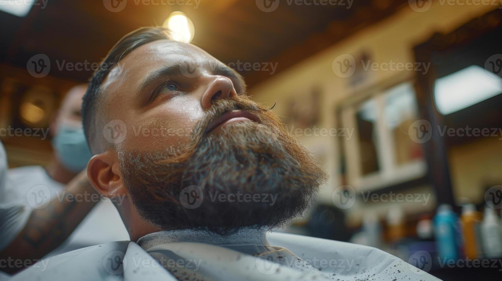 A man with a full beard is getting a trim at a barbershop. He is relaxed in a cape while a barber carefully styles his facial hair. The atmosphere is busy and inviting. photo