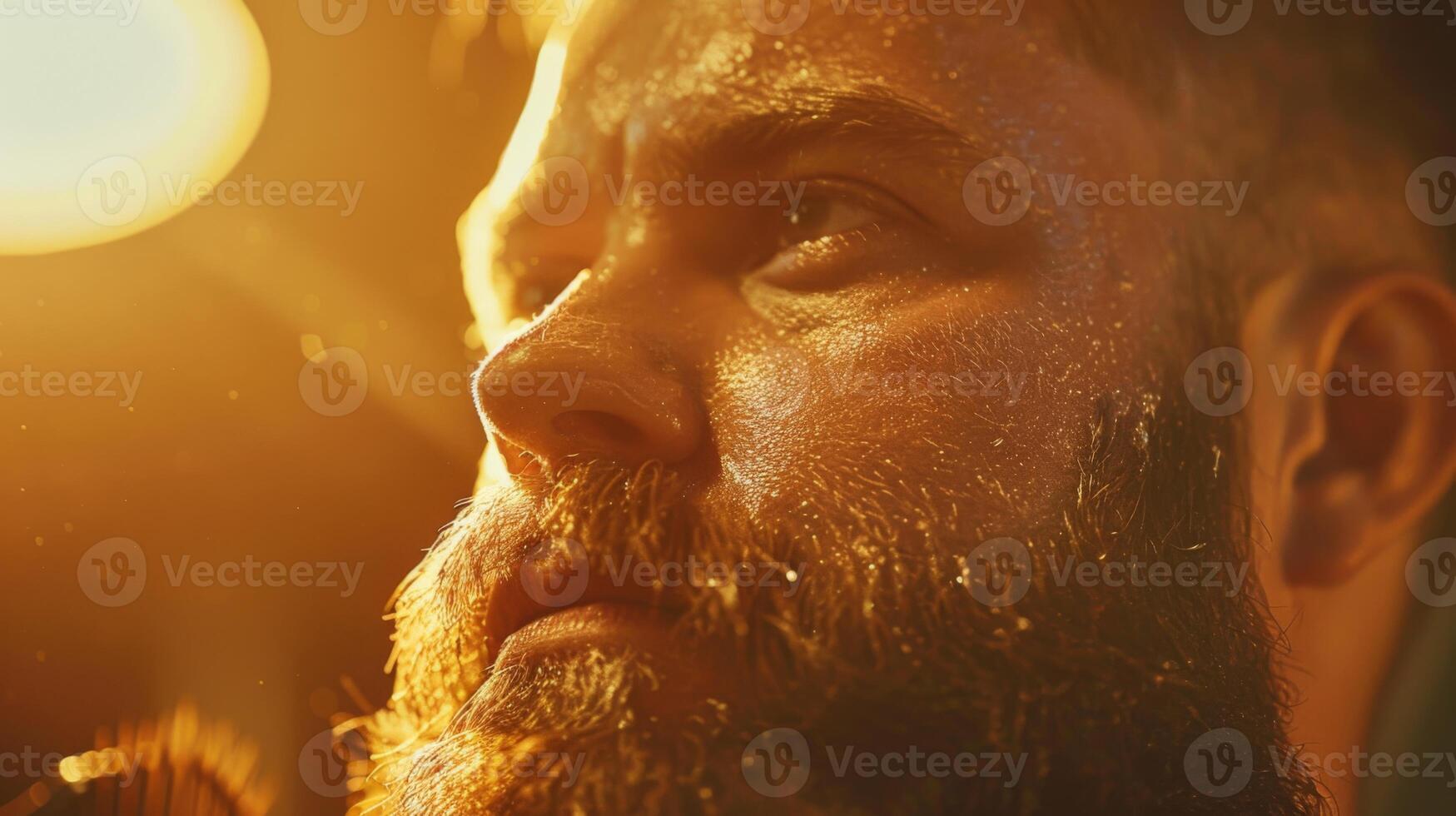 A man with a well groomed beard is captured in a concentrated expression, surrounded by warm lighting that highlights his features in a low lit environment. photo