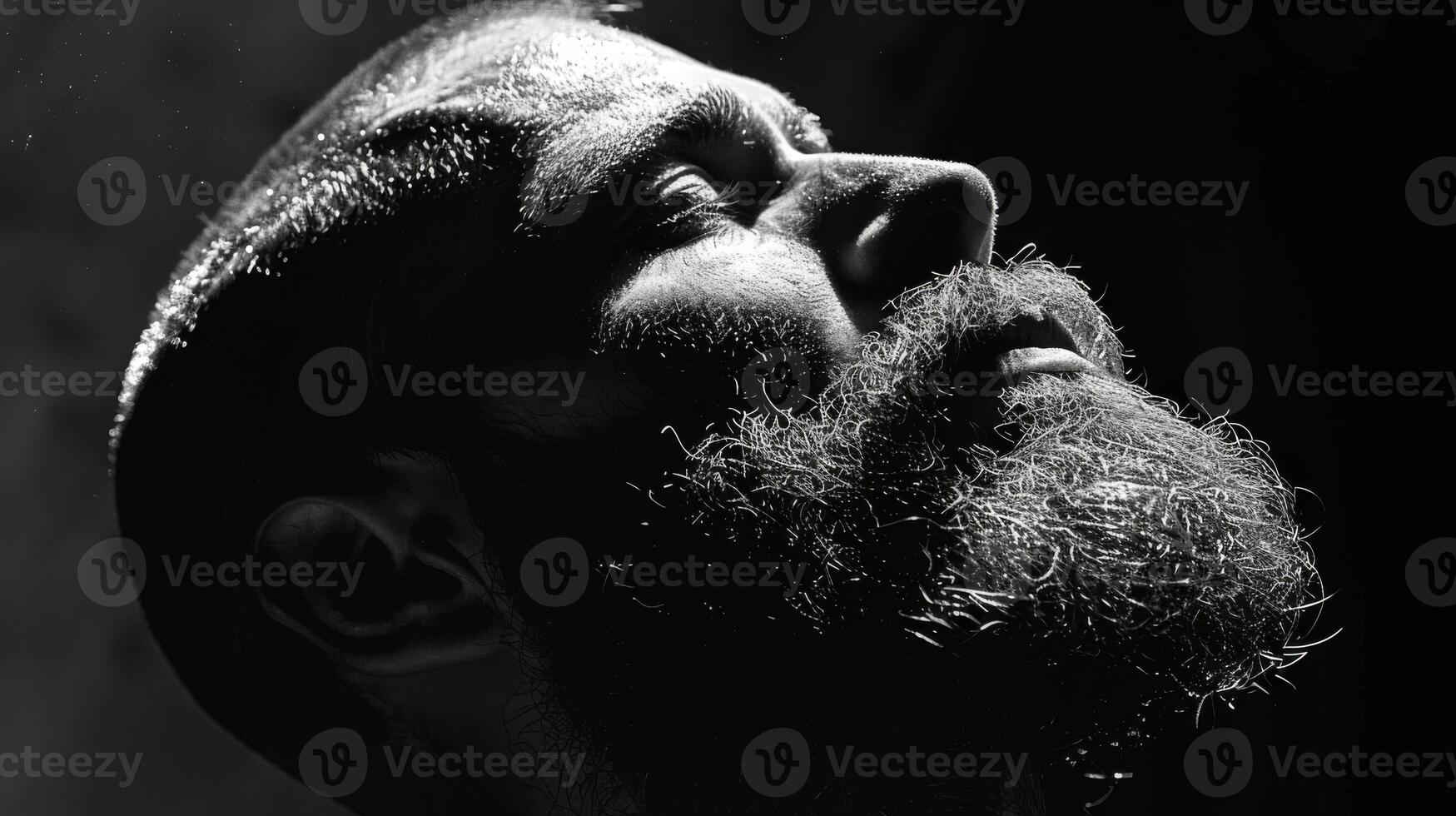 A man with a full beard tilts his head back, evoking a contemplative mood. The interplay of light and shadows creates a striking visual, highlighting facial features. photo