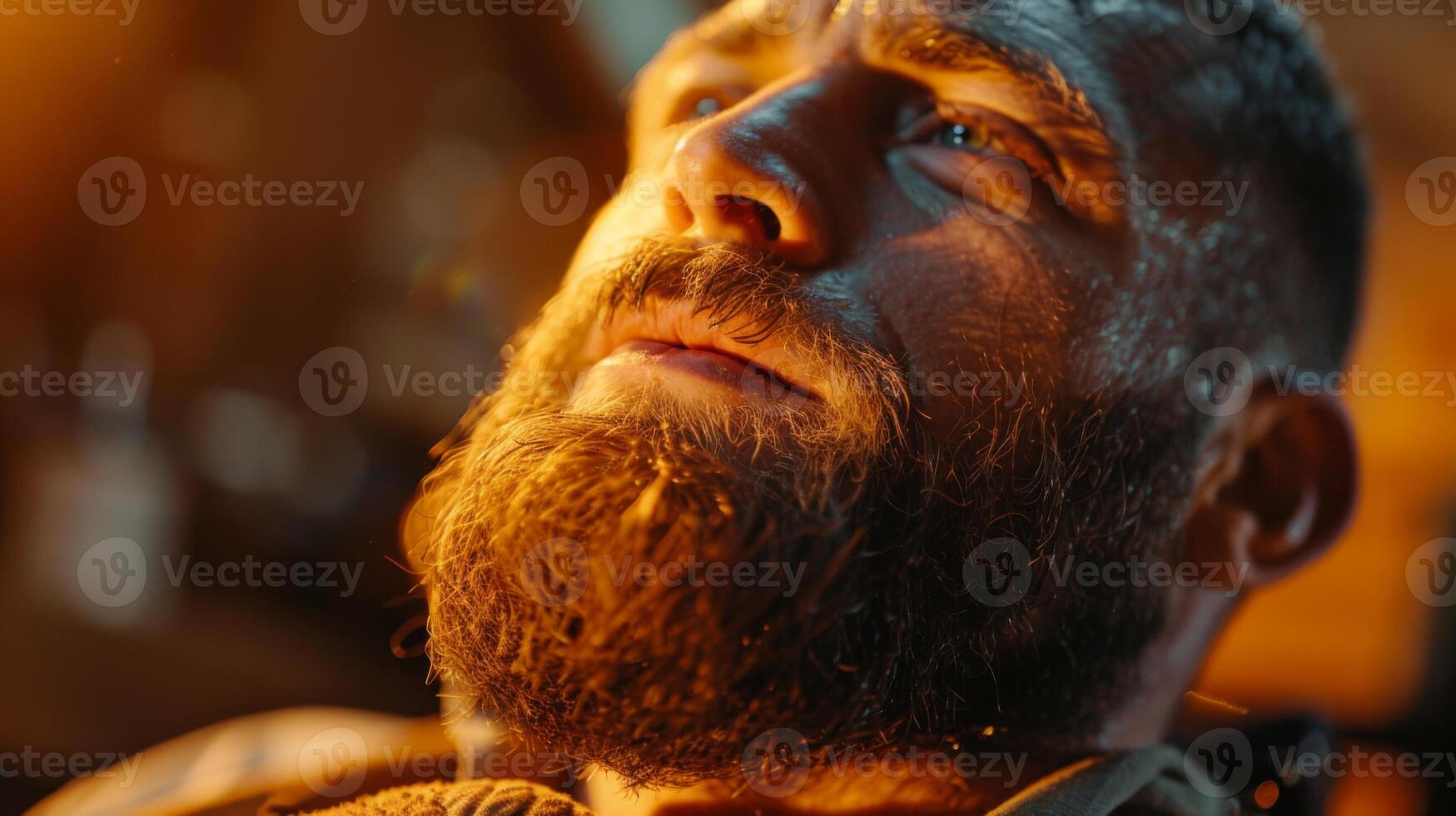A man with a well groomed beard gazes upward, lost in thought, as soft golden light illuminates his face, highlighting the intensity of his expression. photo