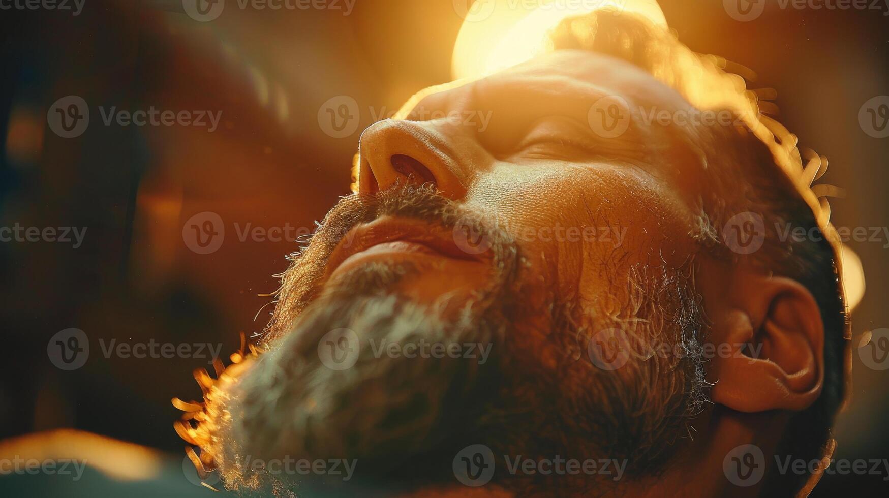 A bearded man relaxes, eyes closed, basking in the warm glow of sunlight. The atmosphere is soothing as he savors a moment of peace and serenity in an inviting indoor space. photo