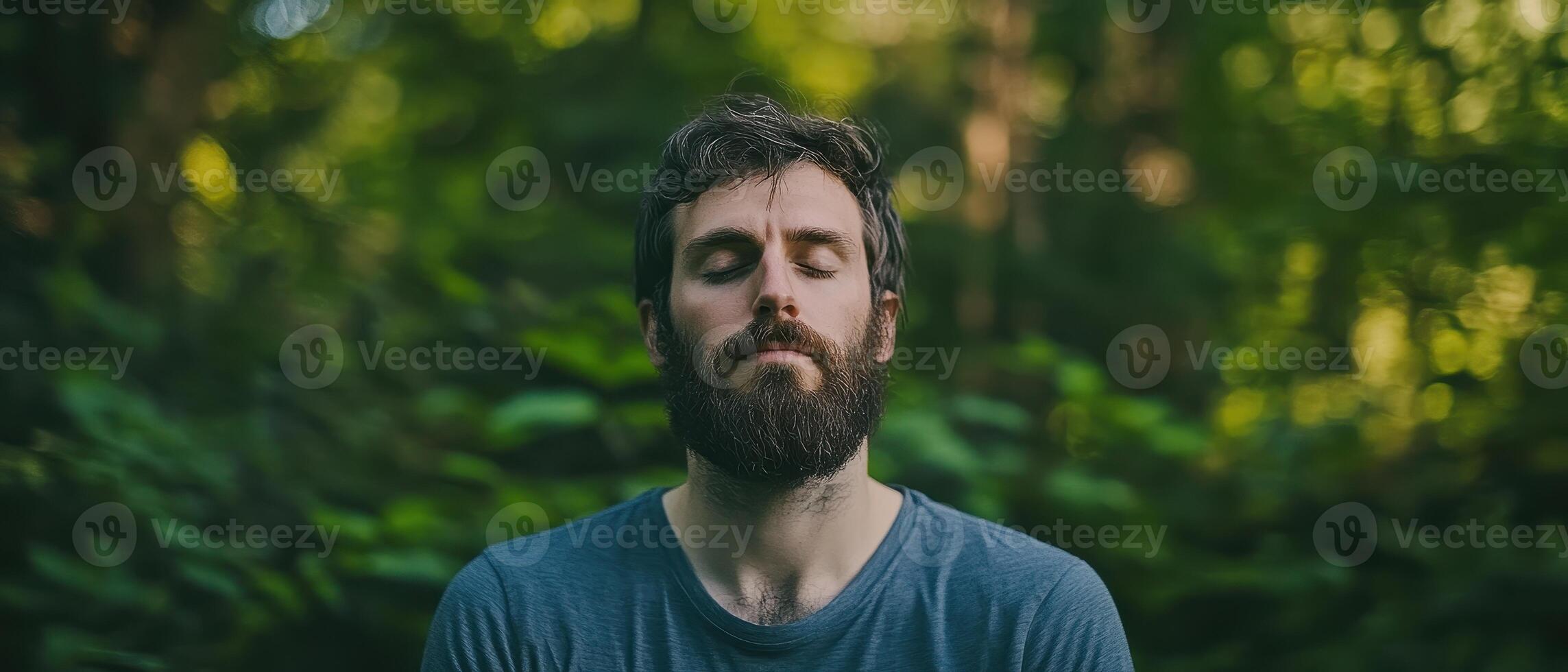 A serene man with a beard meditates in a lush, green forest, embodying tranquility and connection with nature. photo