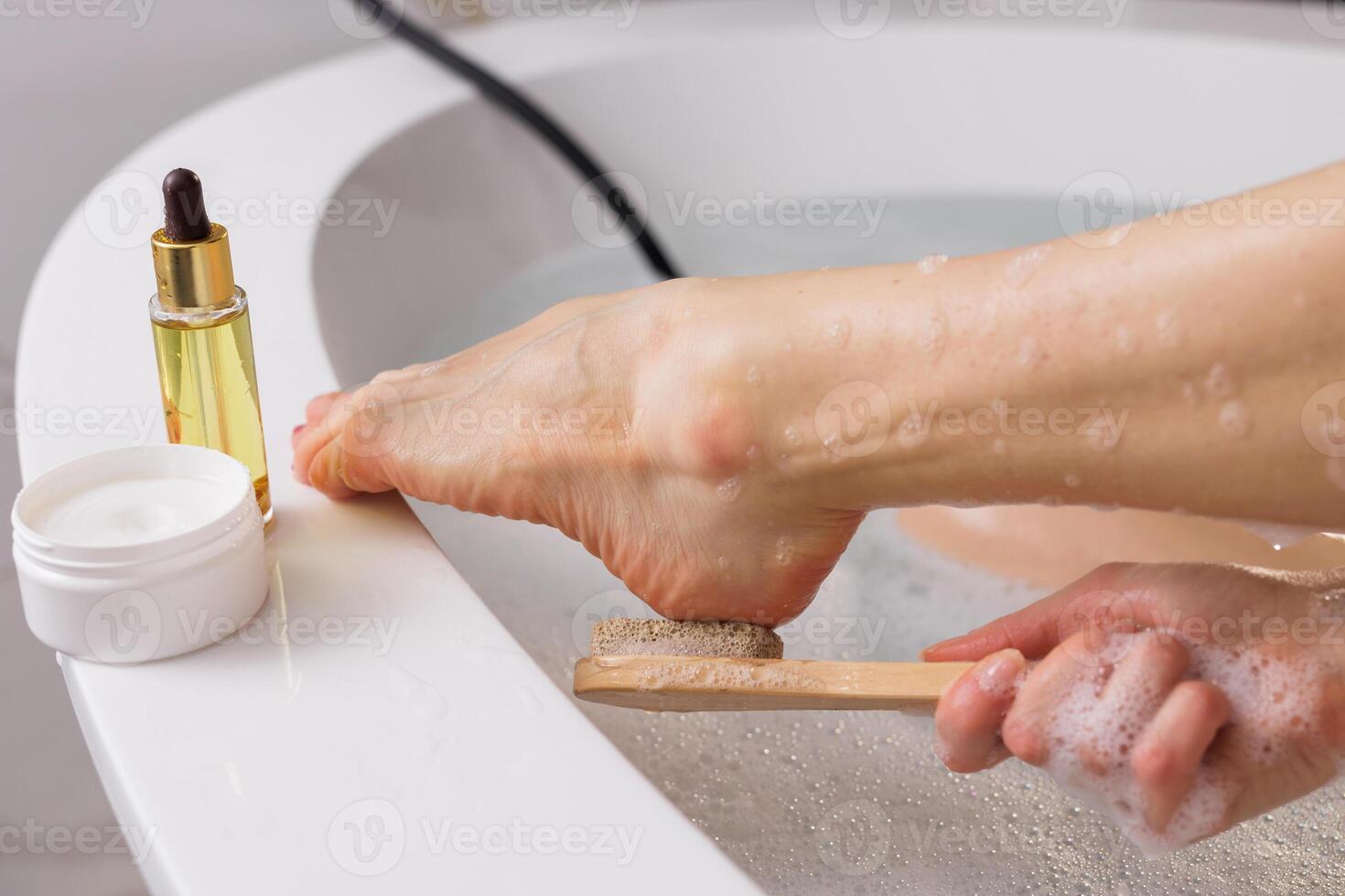 Foot care and relaxation. Woman exfoliating foot with a pumice stone in a warm bath. Skincare essentials like cream and oil nearby. Spa, self-care, and wellness concept photo