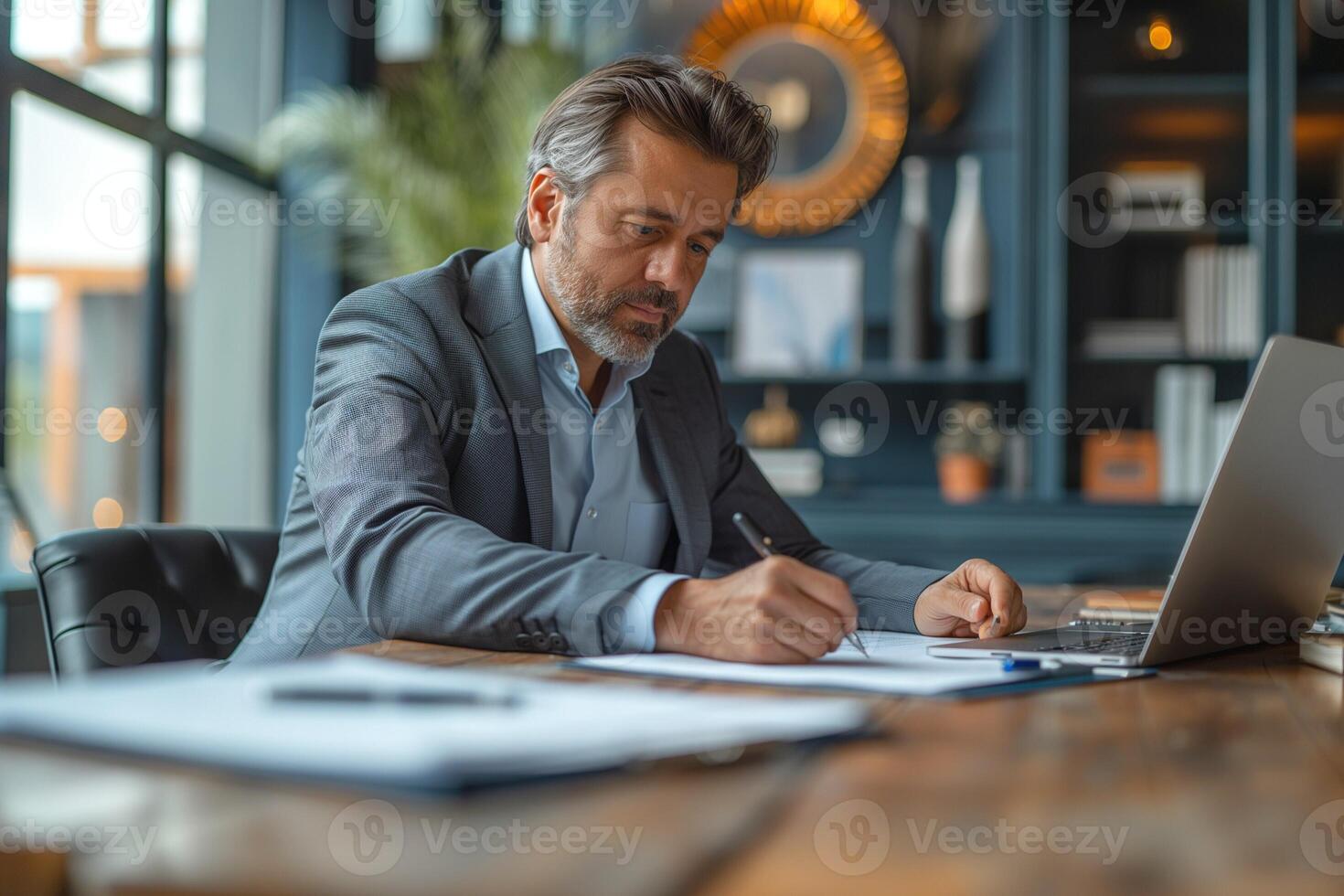 A focused middle-aged Caucasian man in a gray suit writes notes at his desk, with a laptop and modern decor in the background. photo