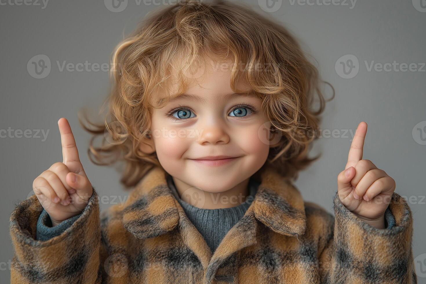 Young boy with curly hair smiles joyfully, raising his fingers in excitement against a soft gray background. photo