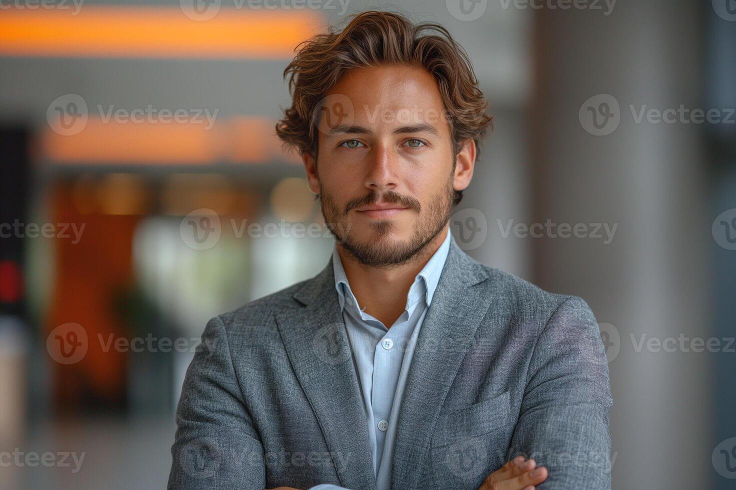 Confident young Caucasian man in a gray blazer, smiling with arms crossed in a modern office setting. photo