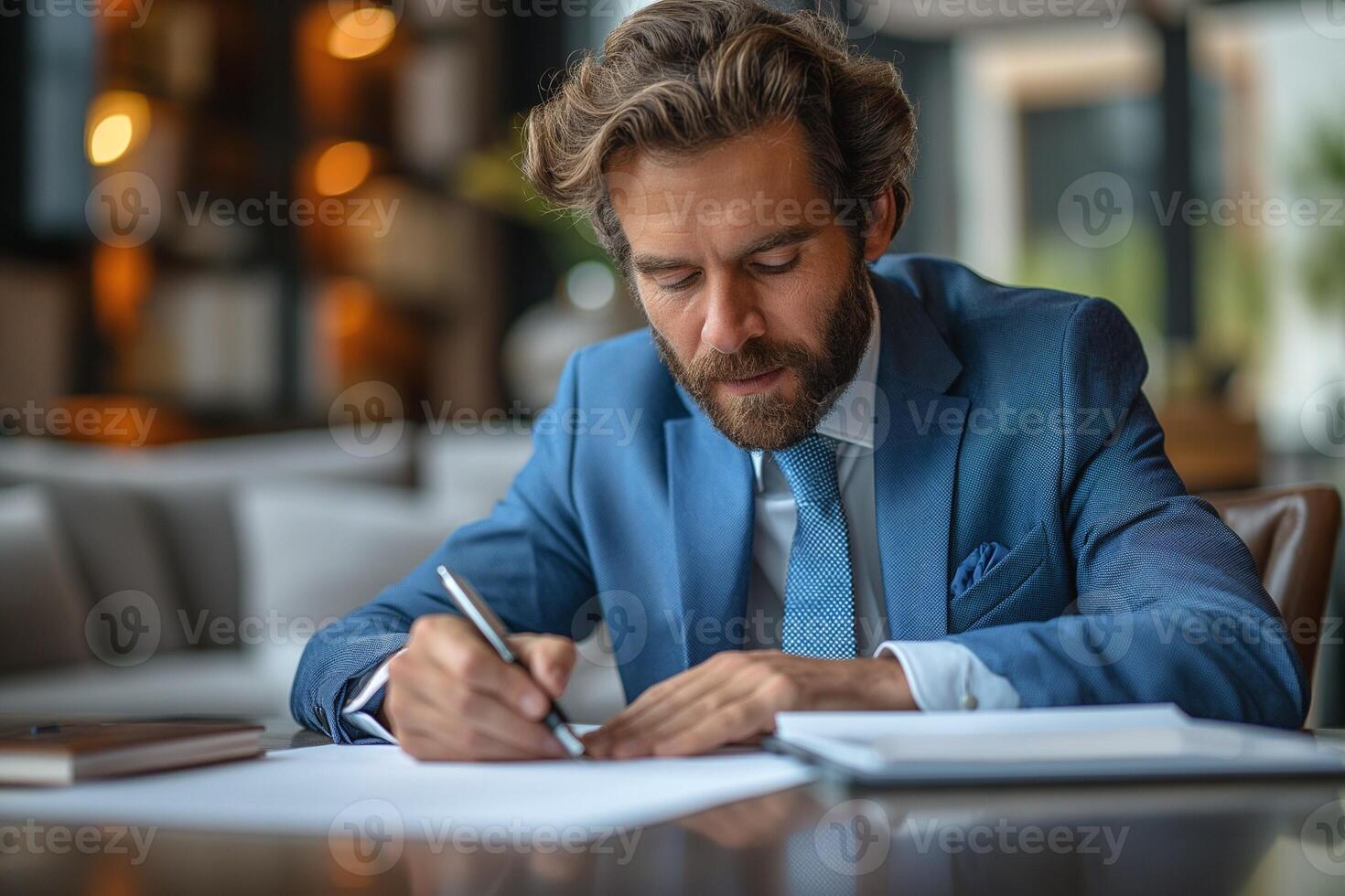 A focused Caucasian man in a blue suit writes notes in a stylish office setting, showcasing professionalism and concentration. photo