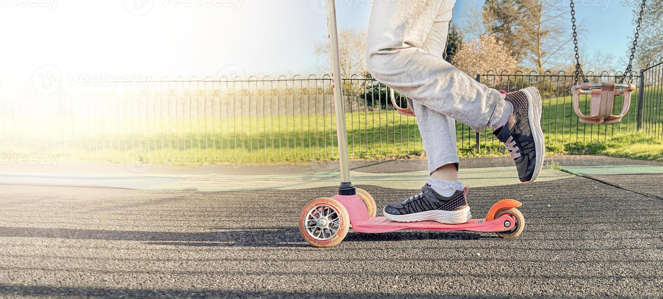 Kid riding a scooter in a park on a playground. Feet wearing sneakers trainers close up. banner photo
