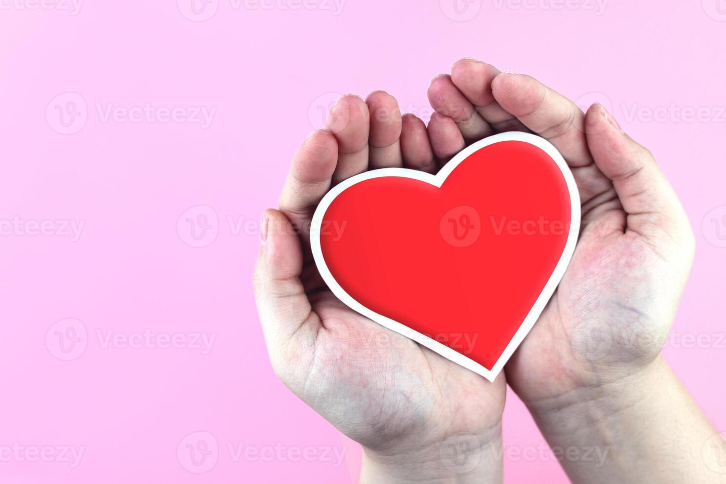 Red paper heart in hand on pink background. Giving love, Valentine's Day, and concept of heart health photo