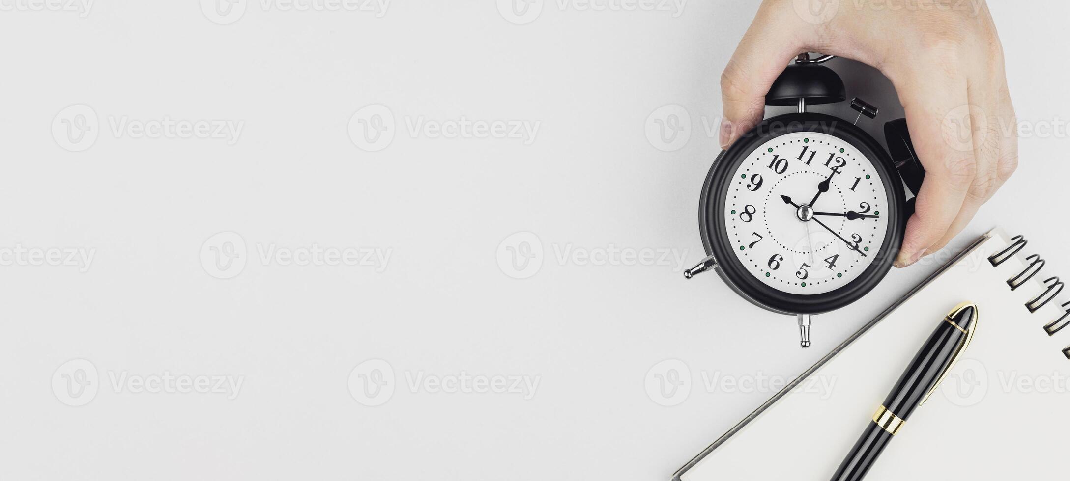 Hand holding a black alarm clock with notebook and pen on white background, with copy space. Concepts of time management, appointment hours, and overtime. photo