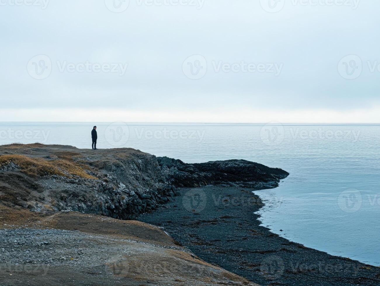 Solitary figure standing on rocky coast with calm waters during overcast afternoon photo