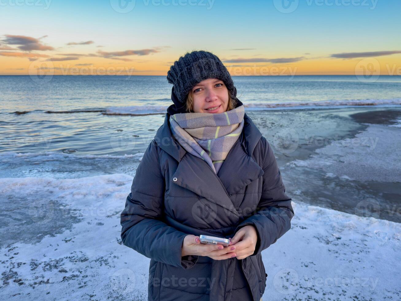 A woman in a warm winter coat, knitted hat, and scarf stands by the frozen shore of Lake Ladoga, smiling gently at the camera. The golden hues of the sunset reflect on the icy water, creating a serene photo
