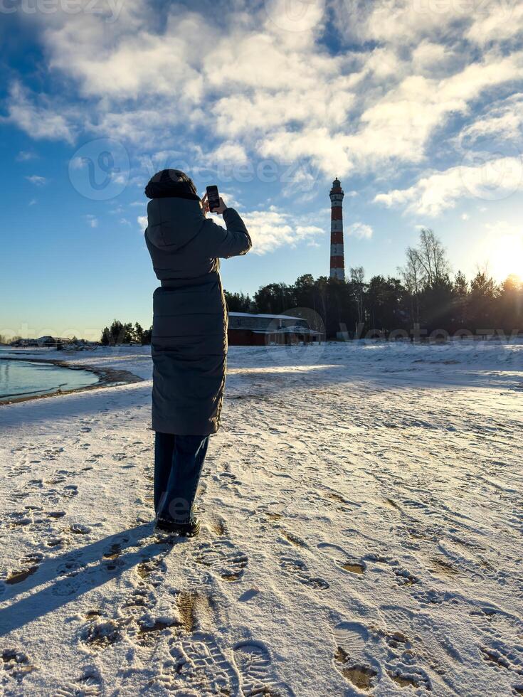 A woman in a long winter coat captures a photo of Osinovets Lighthouse with her smartphone while standing on a snow-covered beach of Lake Ladoga. The setting sun casts long shadows, highlighting the