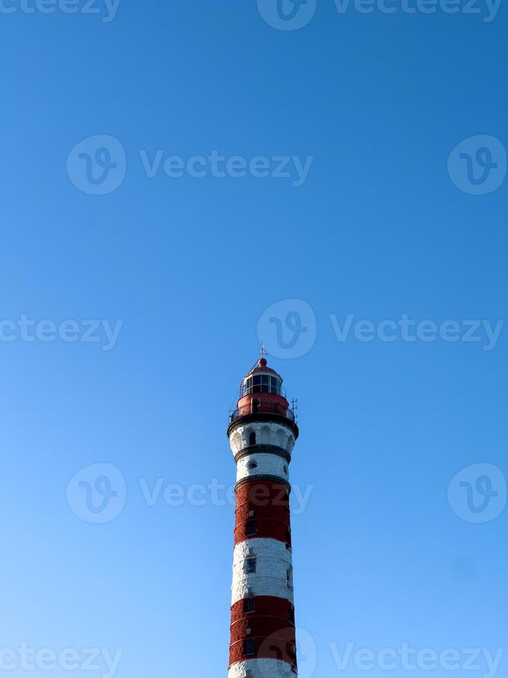 A striking red-and-white Osinovets Lighthouse stands tall against a vibrant blue sky, illuminated by sunlight. The historic maritime beacon, located on the shores of Lake Ladoga, is a significant photo