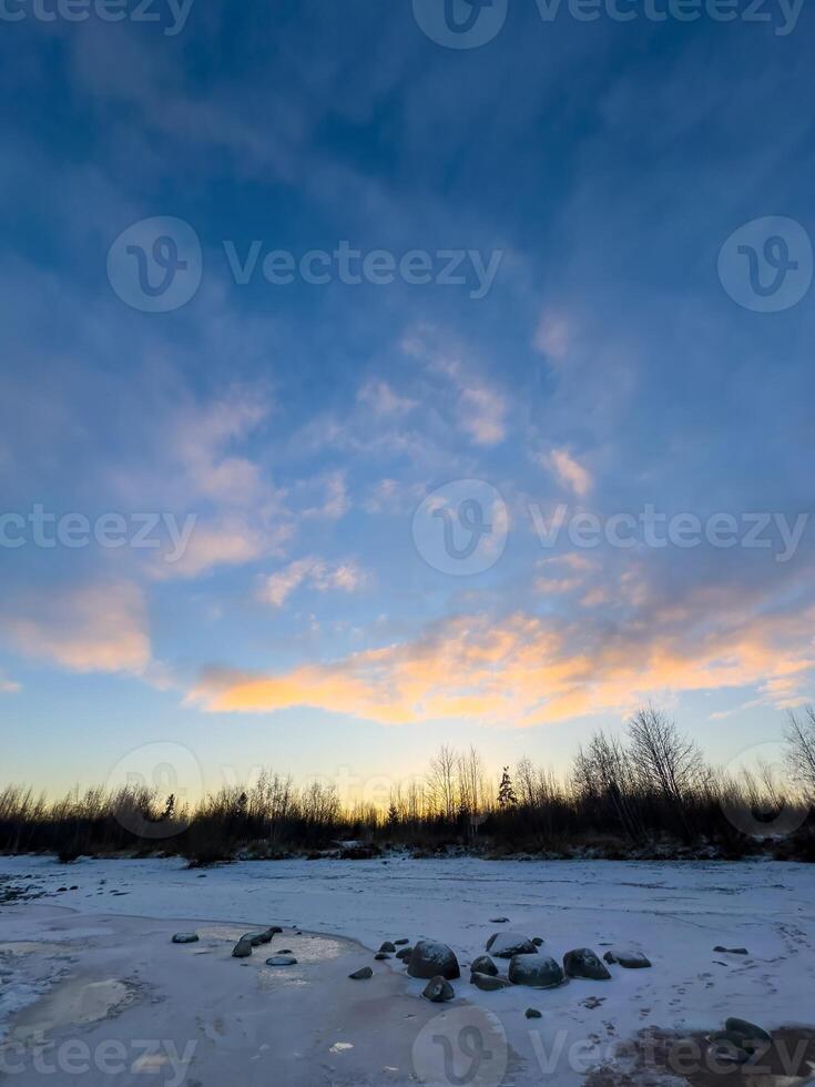 A serene winter sunset paints the sky in shades of blue, orange, and gold over a frozen shoreline. Patches of snow and ice cover the ground, reflecting the soft glow of the setting sun. Bare trees photo