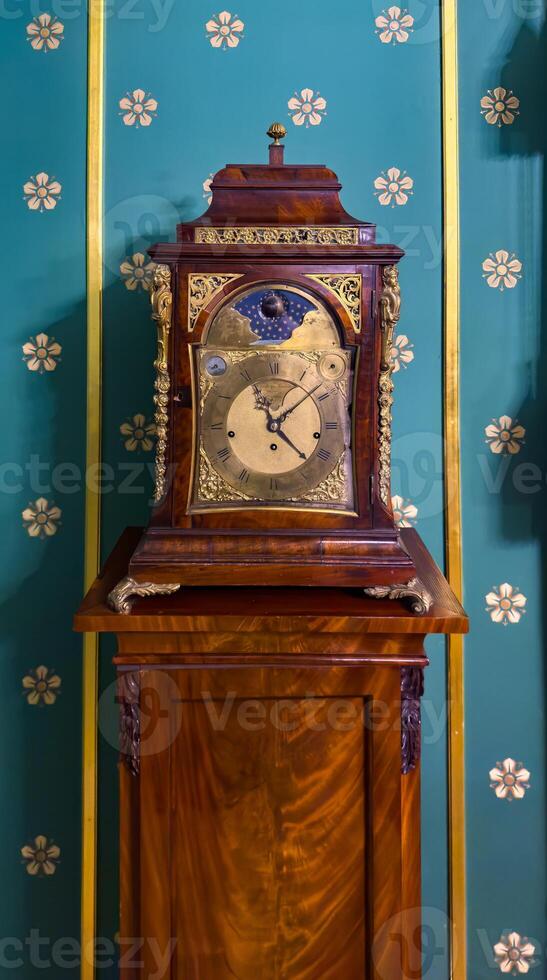 A close-up view of a vintage wooden clock with intricate brass details, set against a rich teal wall adorned with floral patterns. The clocks elegant design reflects the craftsmanship of a bygone era photo
