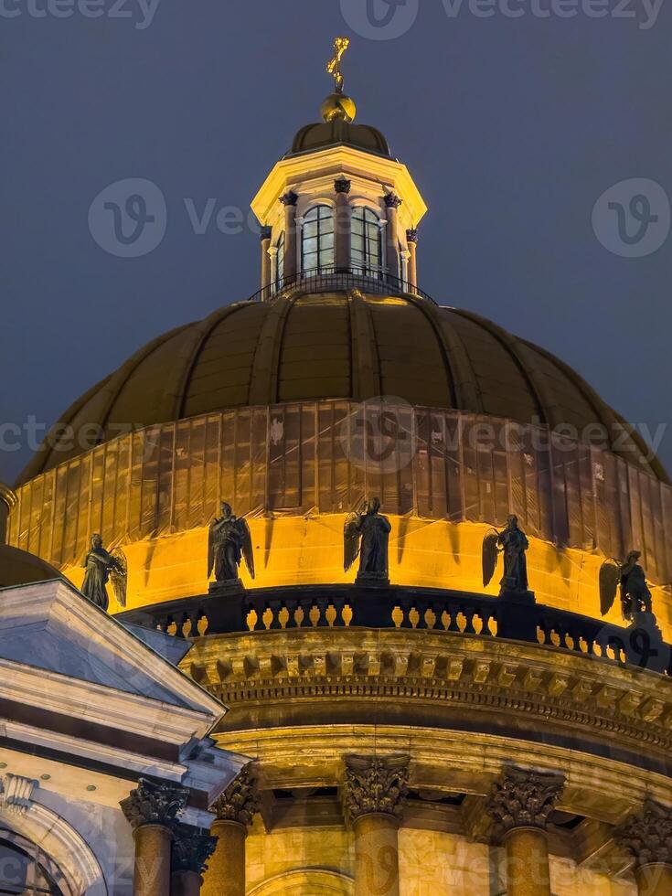 A close-up view of the majestic dome of St. Isaac Cathedral in Saint Petersburg, glowing under golden illumination against the dark evening sky, highlighting its architectural grandeur. photo
