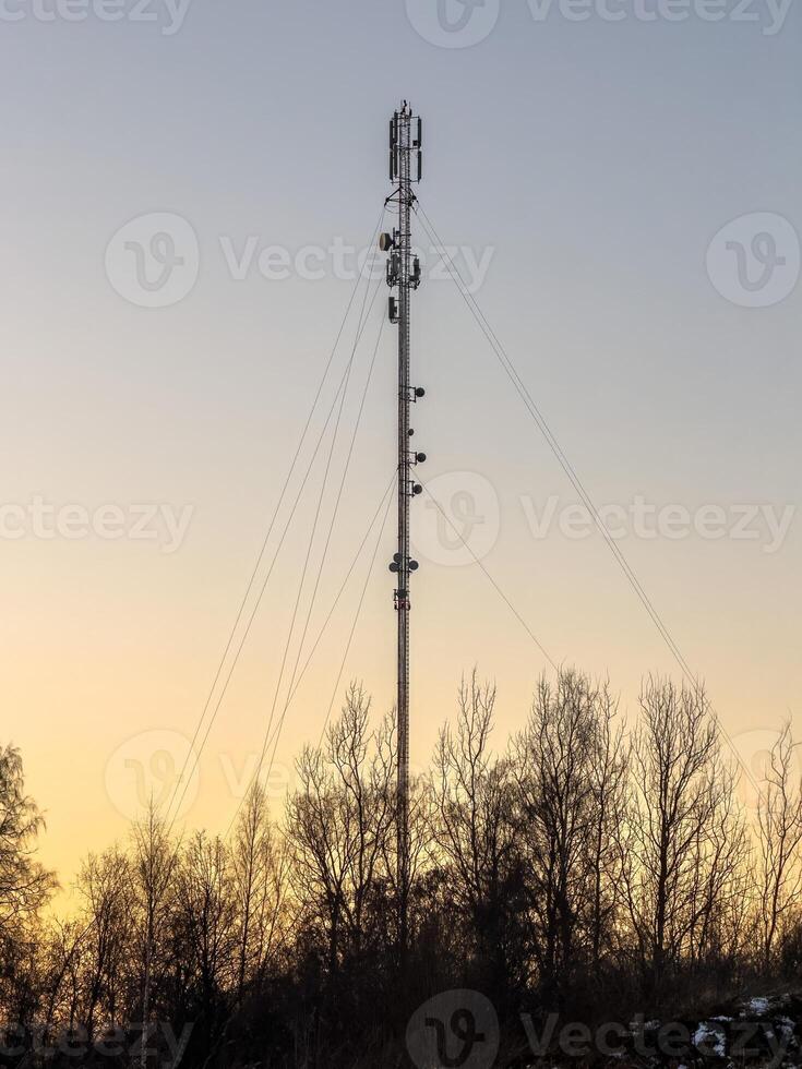 A tall communication tower stands silhouetted against a warm sunset sky, with sparse trees in the foreground. The tower is equipped with various antennas and equipment, emphasizing modern technology photo