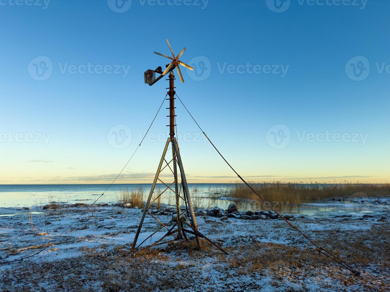 An old windmill with wooden blades stands on the snowy shore of Lake Ladoga, with clear skies and distant reeds lining the horizon. The tranquil winter landscape captures the beauty of the northern photo
