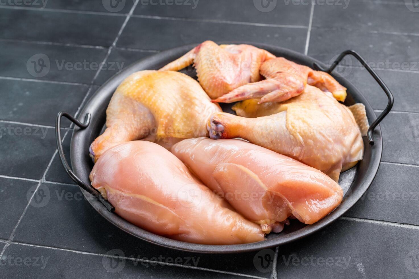 A black metal dish with a handle holds several pieces of fresh, raw chicken. The chicken is arranged neatly in the dish, and the background is a dark, tiled surface. photo
