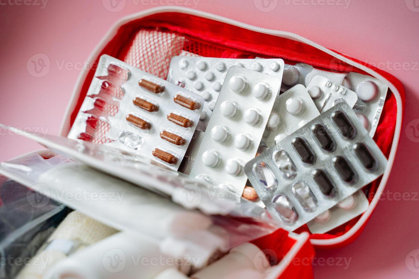 On a soft pink background, there is a vibrant red first aid kit that is generously filled with various pills, capsules, and bandages, ensuring preparedness for any medical need photo