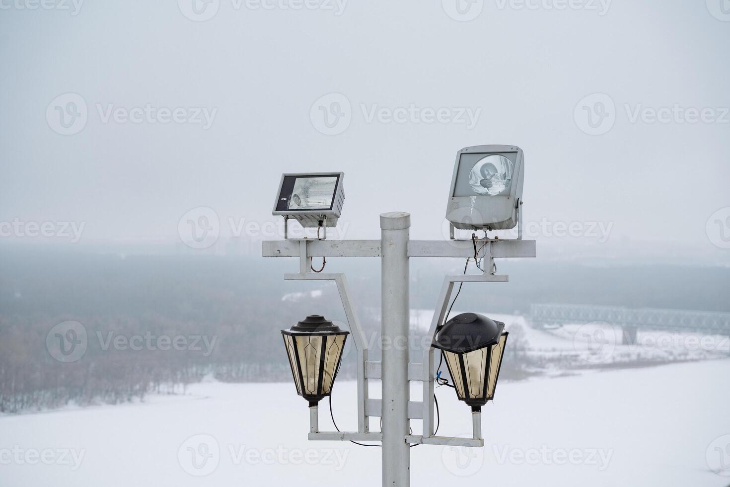 On a chilly winter day, two elegant street lights on a sturdy pole emit a warm glow, as soft snowflakes drift from the sky, enhancing the serene beauty of the white landscape photo