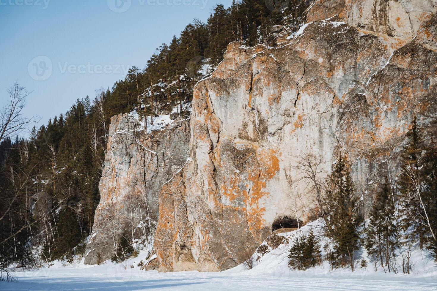 A significant and large rock formation that features a cave nestled within it is completely surrounded by a blanket of snow and tall trees, creating a picturesque winter landscape photo