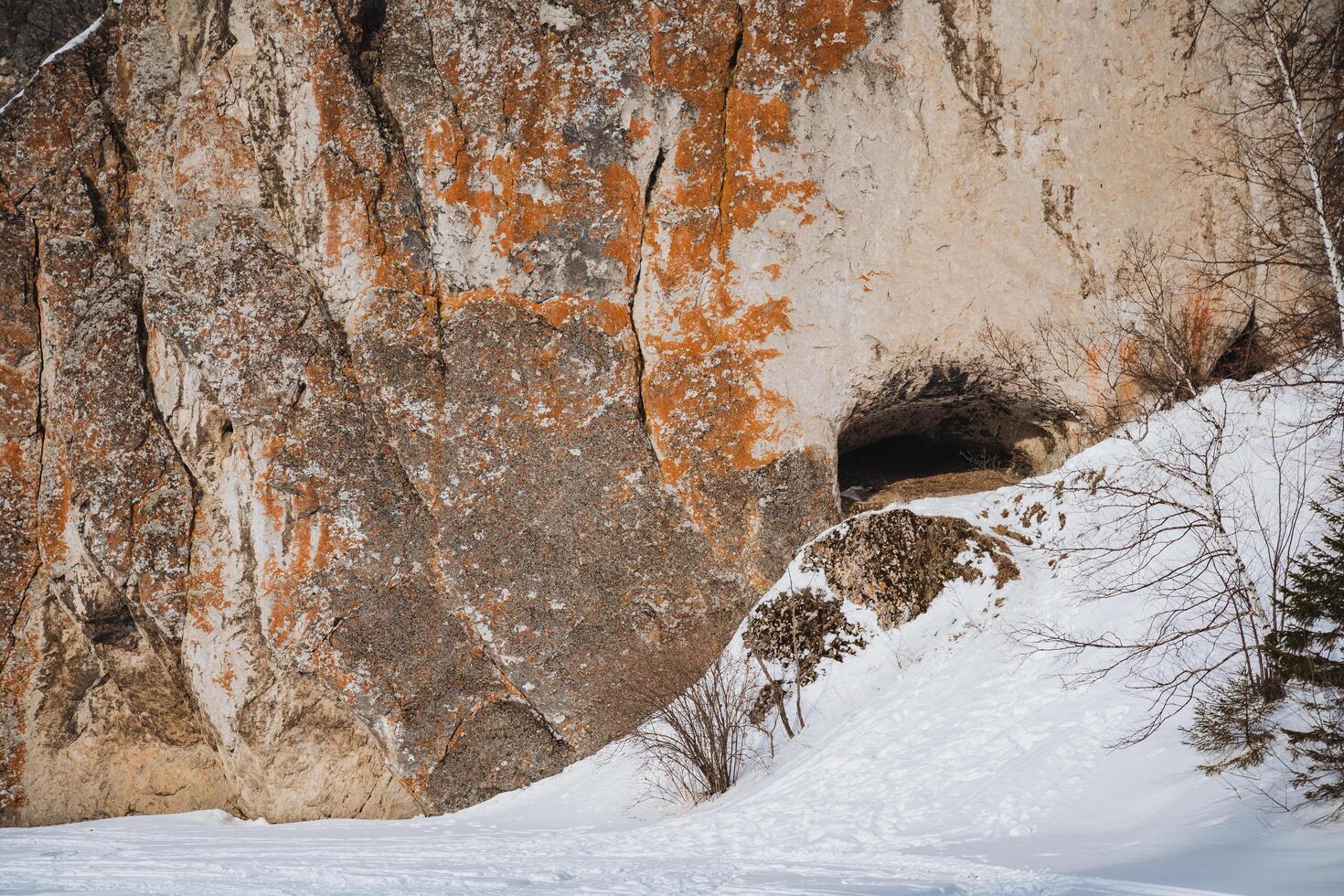 There is a cave nestled in the side of a majestic mountain, which is beautifully surrounded by a blanket of pure white snow and rugged rocks that add to its stunning appearance photo