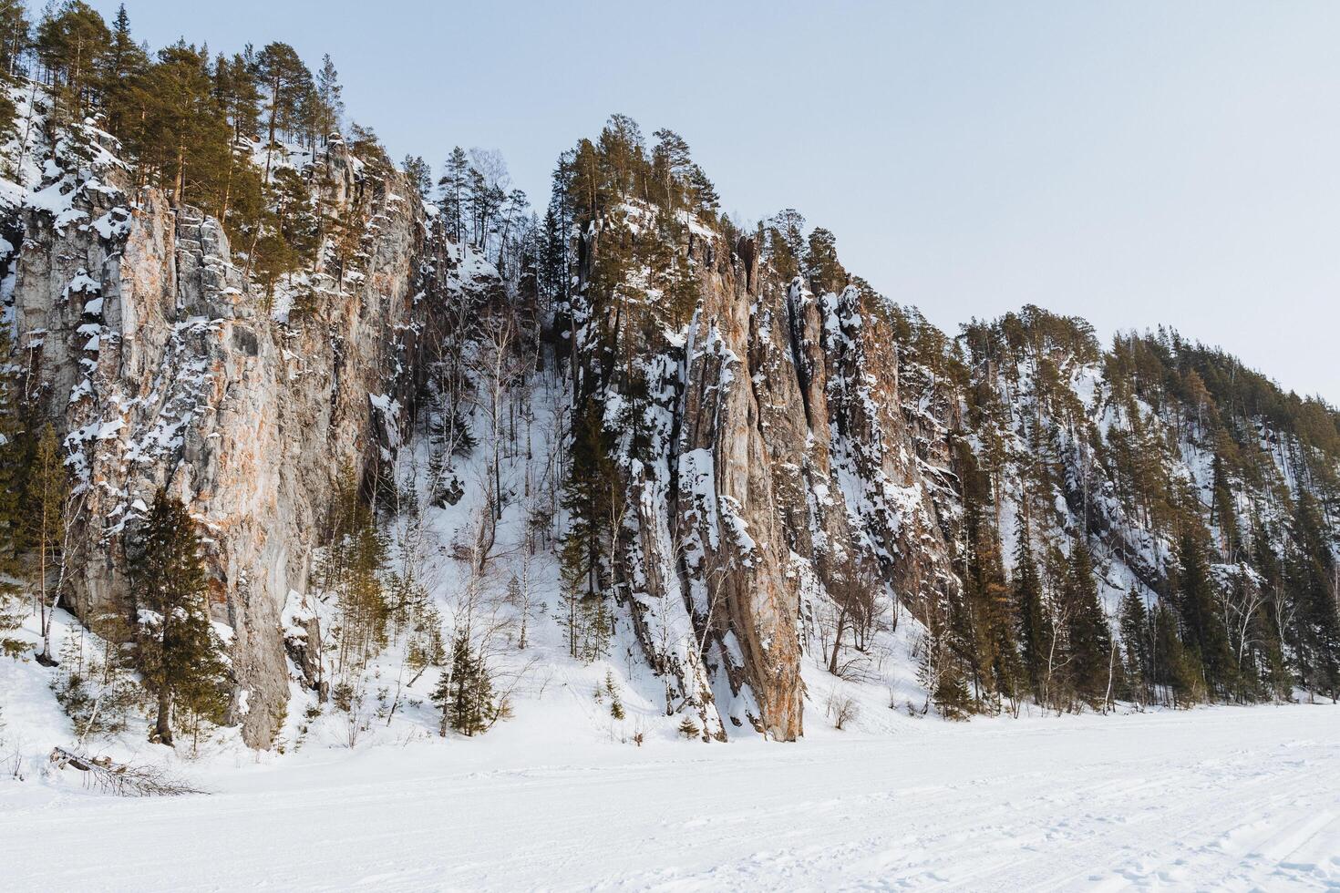 In the picturesque scene, a snowy mountain adorned with a blanket of trees and rugged rocks stands majestically, while a serene, frozen lake glistens invitingly in the foreground photo
