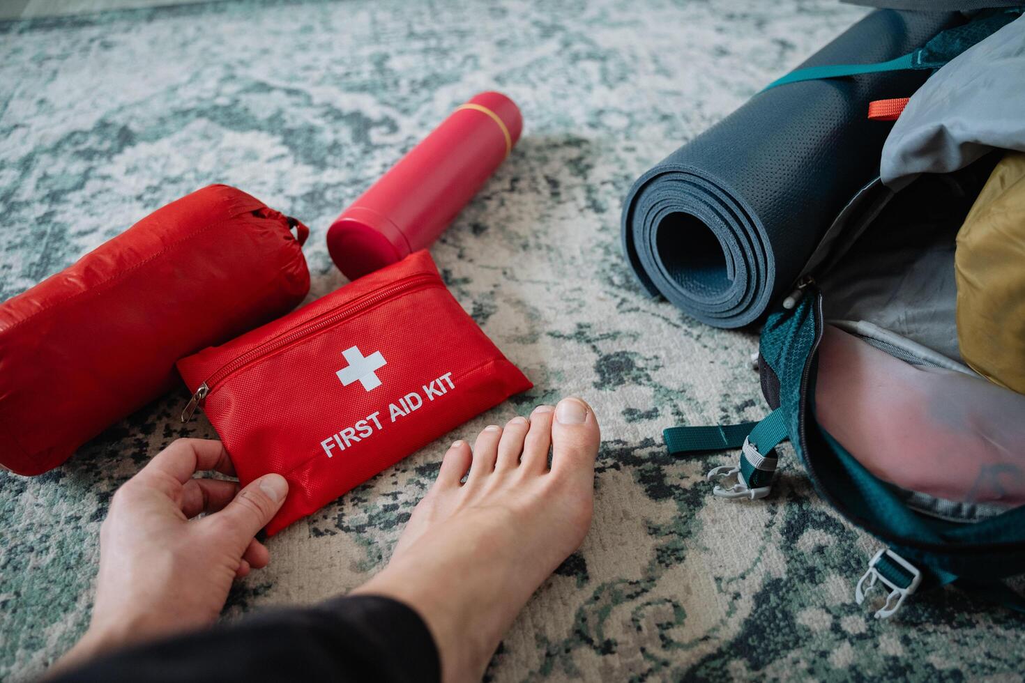 A person is holding a first aid kit in close proximity to a backpack, which could contain various items for outdoor activities or travel purposes like sleeping pads and gear photo