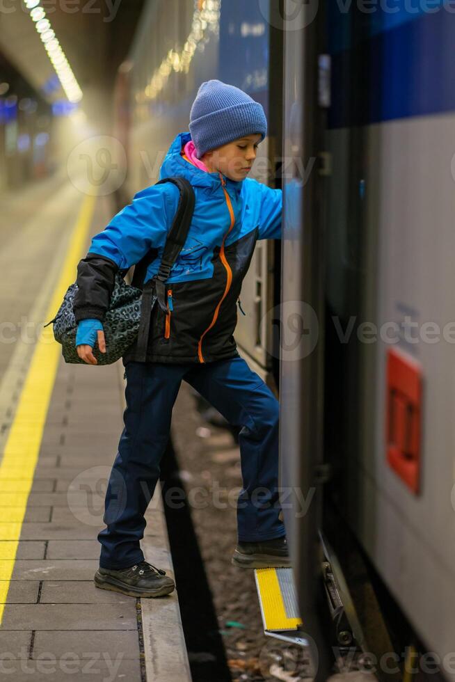 A schoolboy in a blue jacket and hat with a backpack enters a train carriage photo