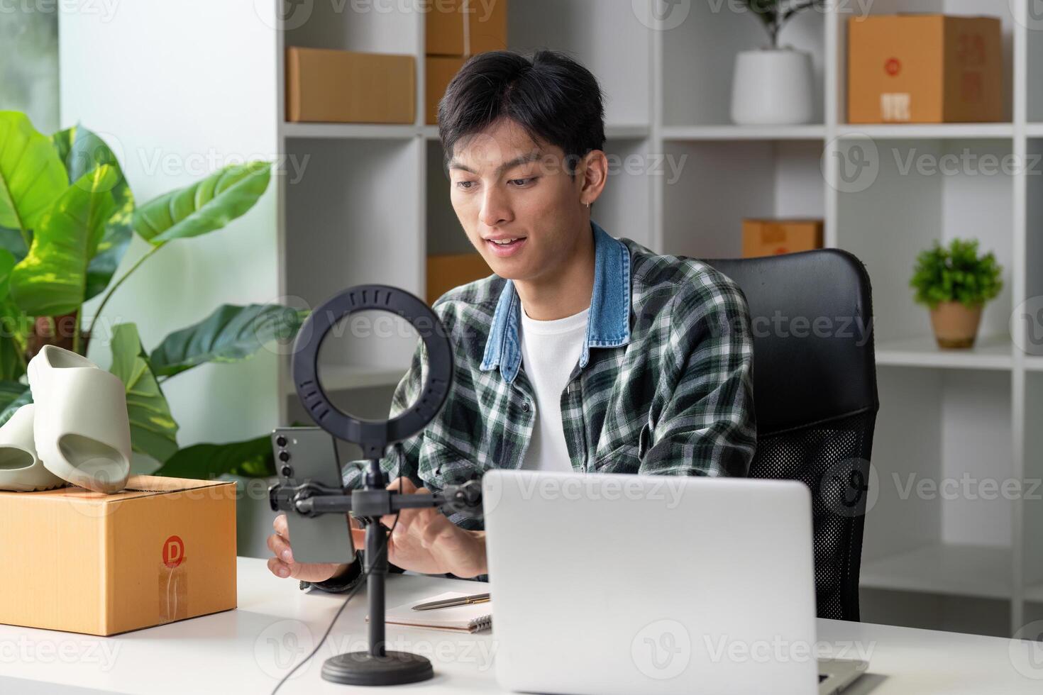 Young entrepreneur selling products online, using smartphone and laptop in modern workspace with greenery. photo