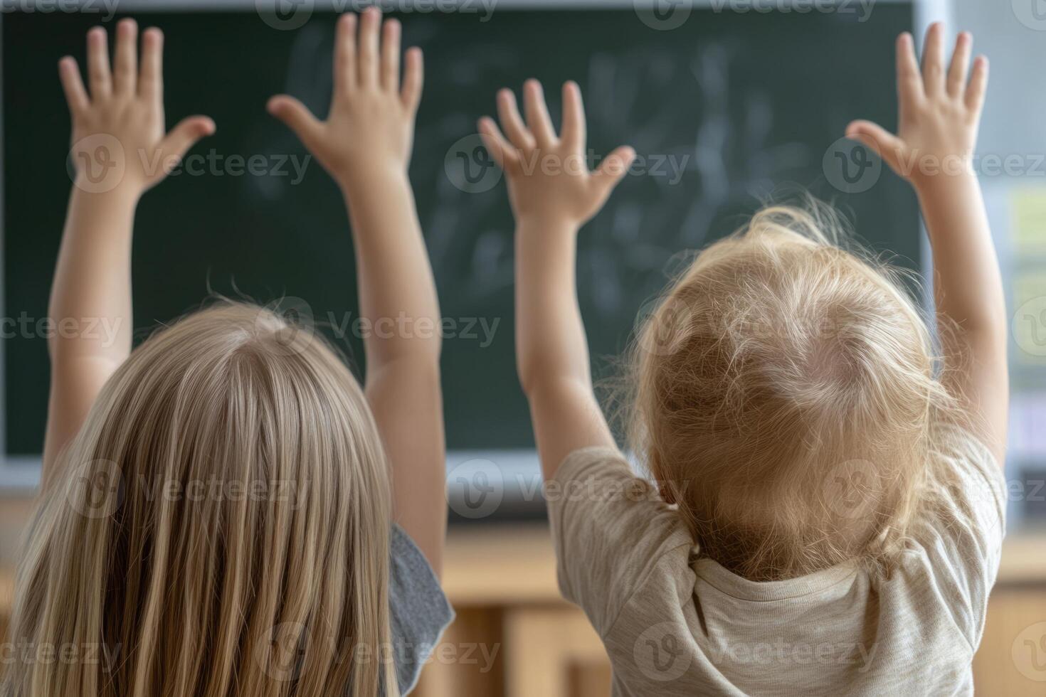 Excited children are raising their hands high in a classroom filled with natural light. Their enthusiasm reflects the joy of learning. The chalkboard displays writing, indicating an engaging lesson photo