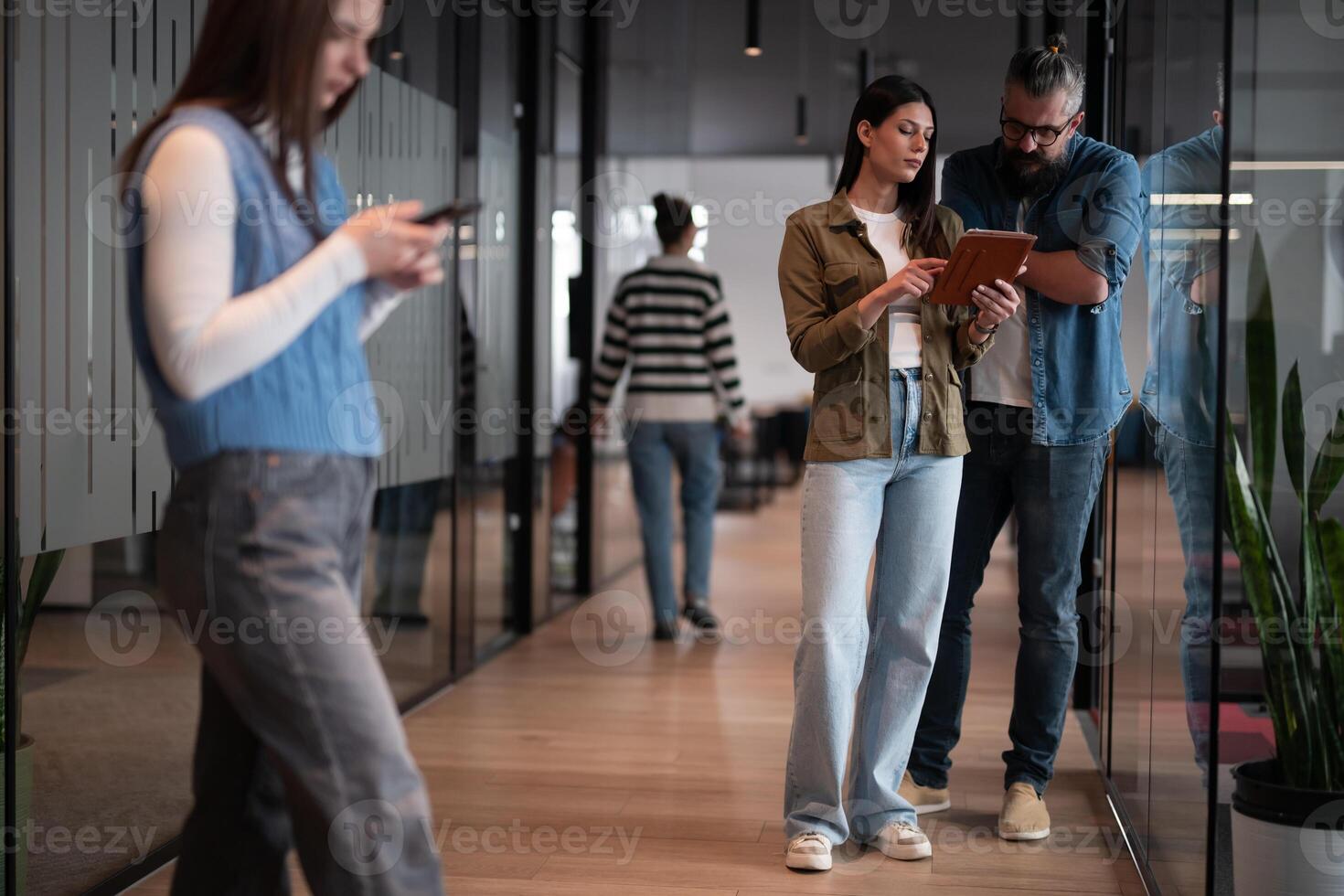 A senior hipster mentor and a European leader discuss a project while working on a tablet in a modern coworking space, collaborating confidently while busy colleagues work in the background photo