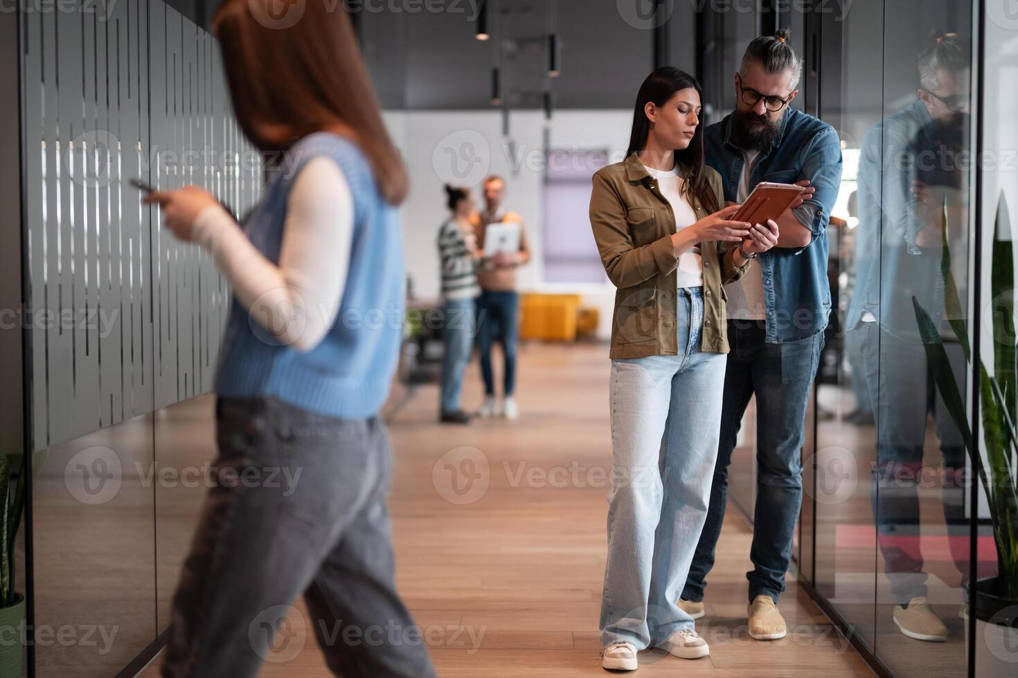A senior hipster mentor and a European leader discuss a project while working on a tablet in a modern coworking space, collaborating confidently while busy colleagues work in the background photo