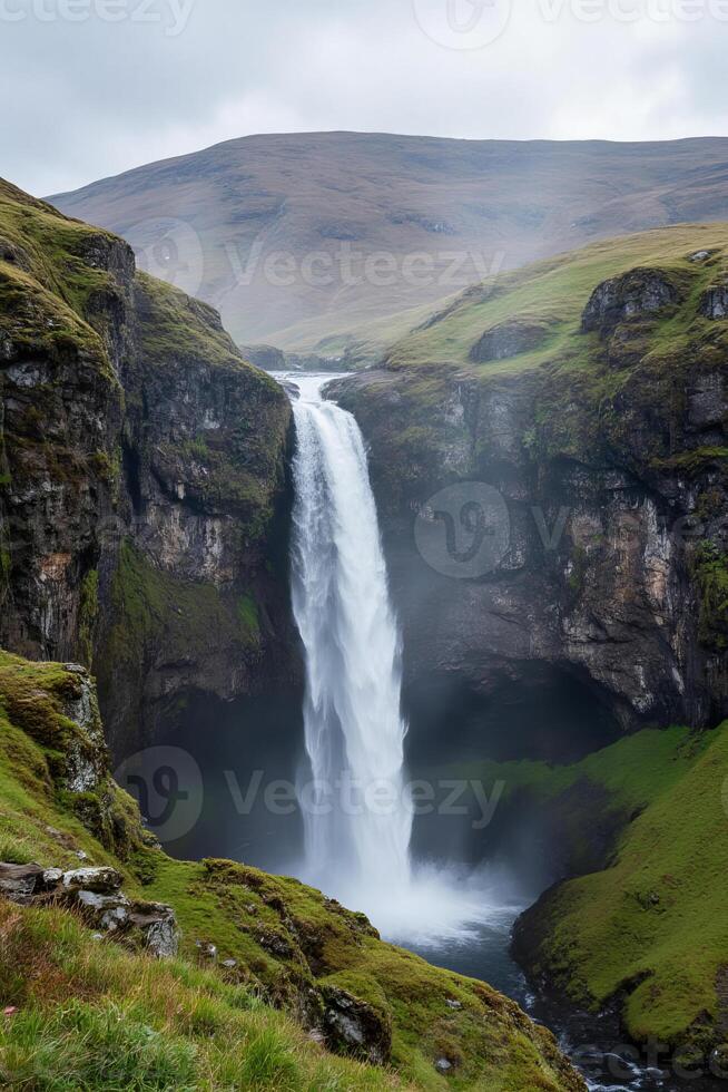 A cascading waterfall tumbles down a rugged cliffside covered in moss, surrounded by the rolling hills photo