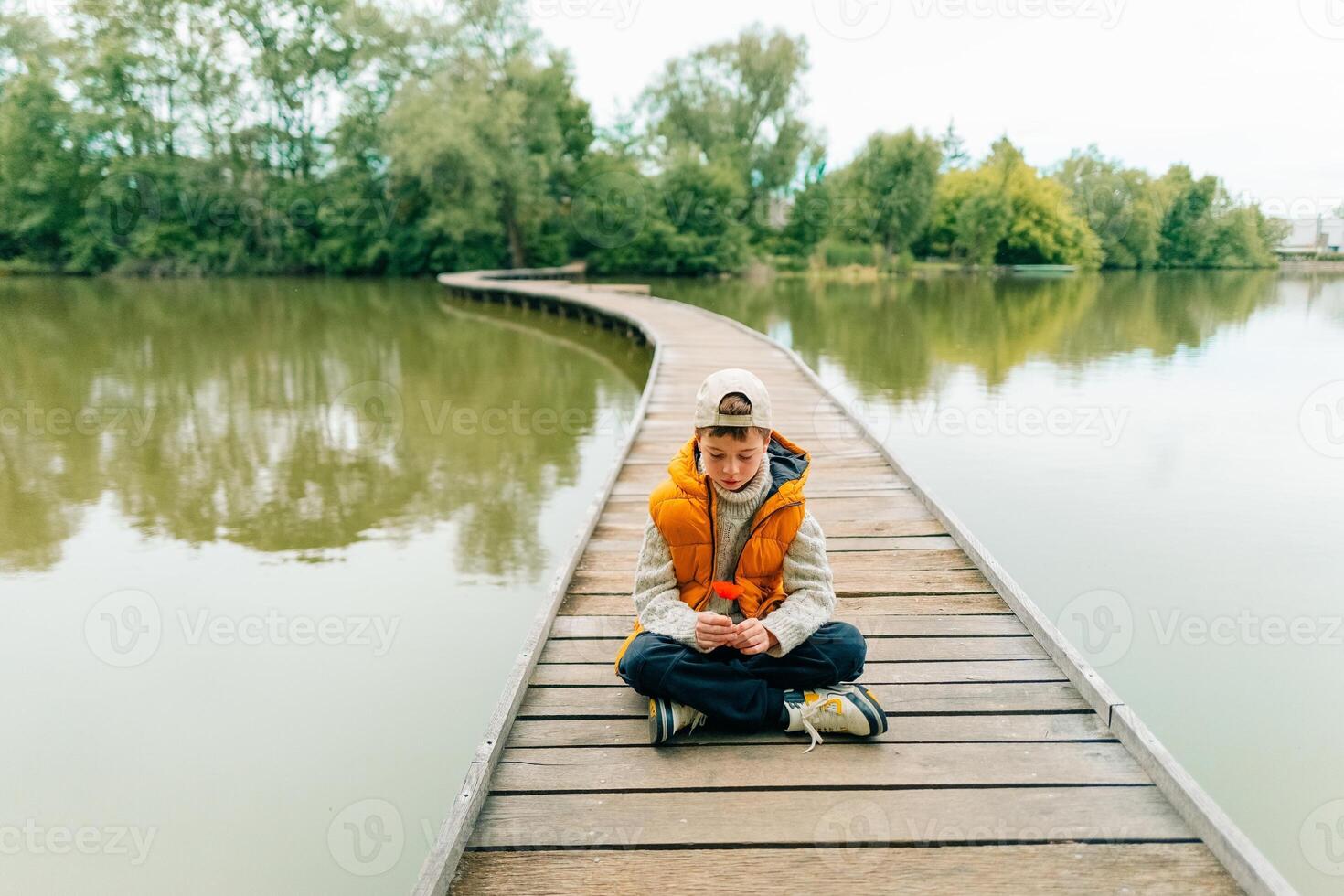 A teenage boy sits on a bridge on a pond and holds a poppy flower in his hands photo