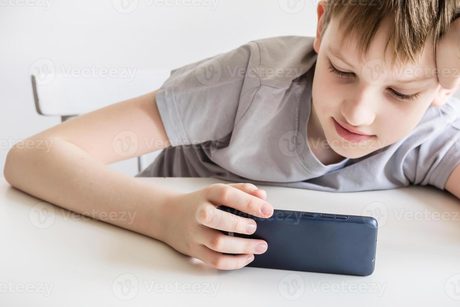 Teen boy sits at white table and uses smartphone photo