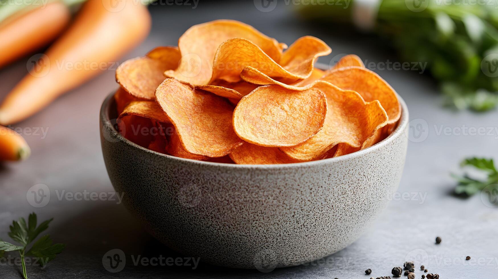 Sweet chips served in a textured bowl with fresh carrots on a dark stone surface and herbs in the background photo