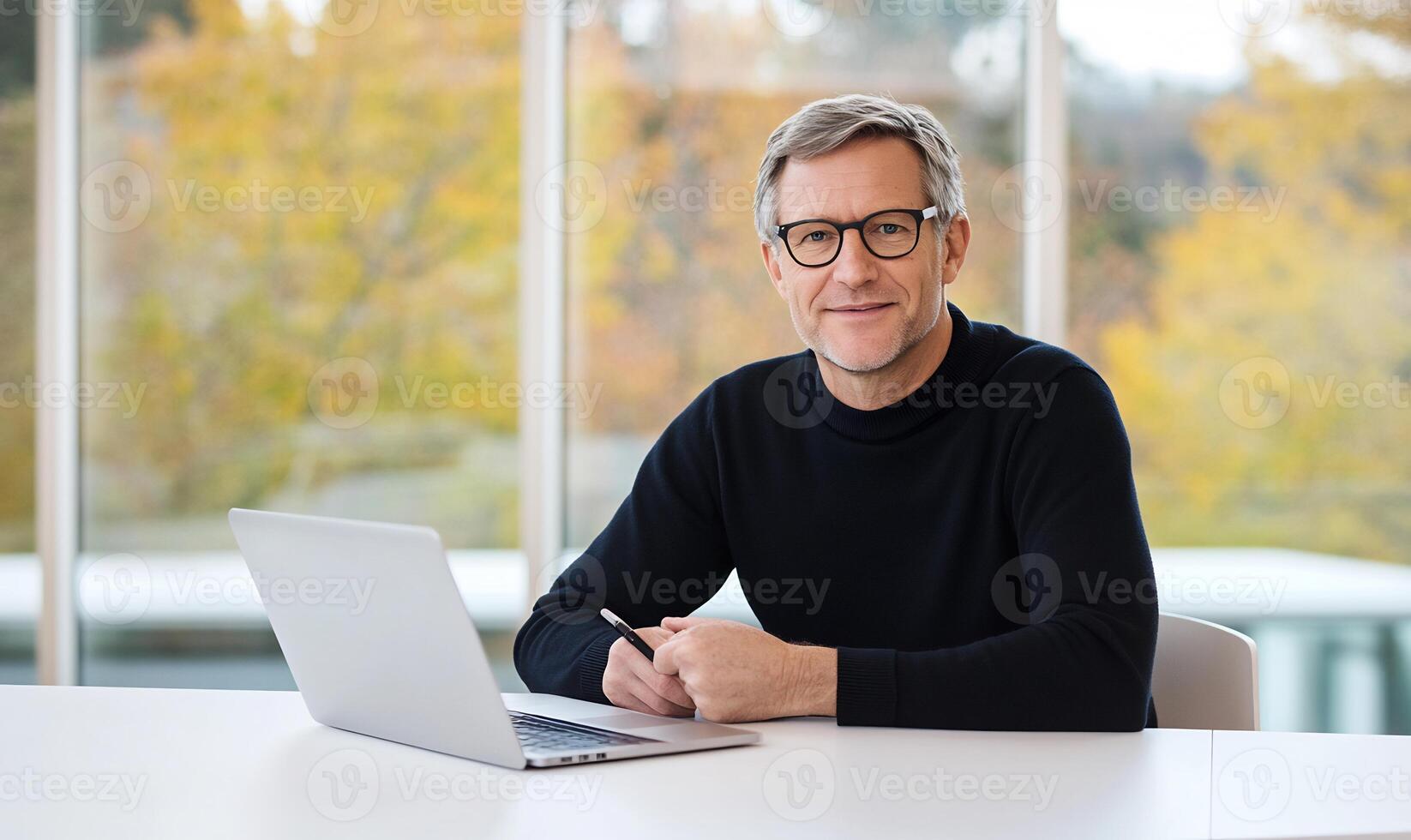 Smiling mature man sits at desk with laptop, pen, and autumn view. photo