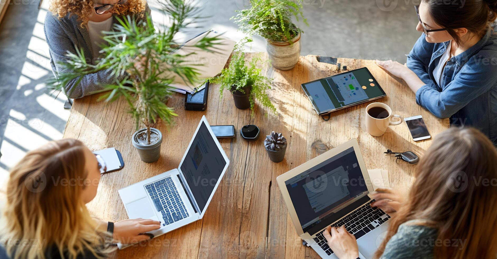 Women collaborating around a wooden table, surrounded by plants and technology, working on laptops and digital tablets. photo