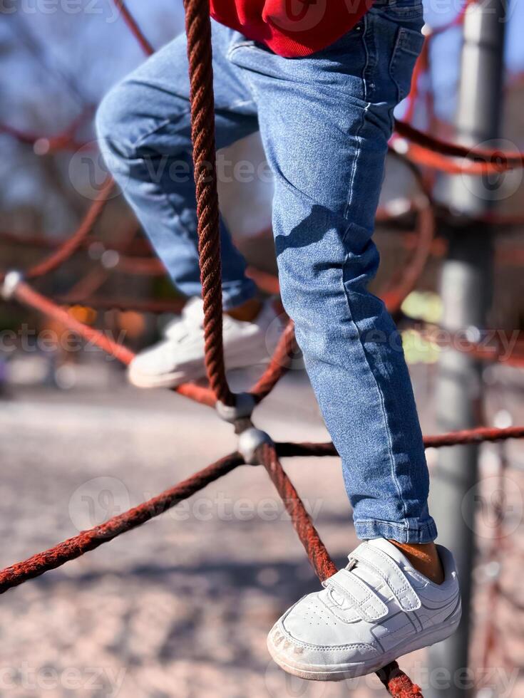 Child climbing rope structure in playground with jeans and sneakers photo