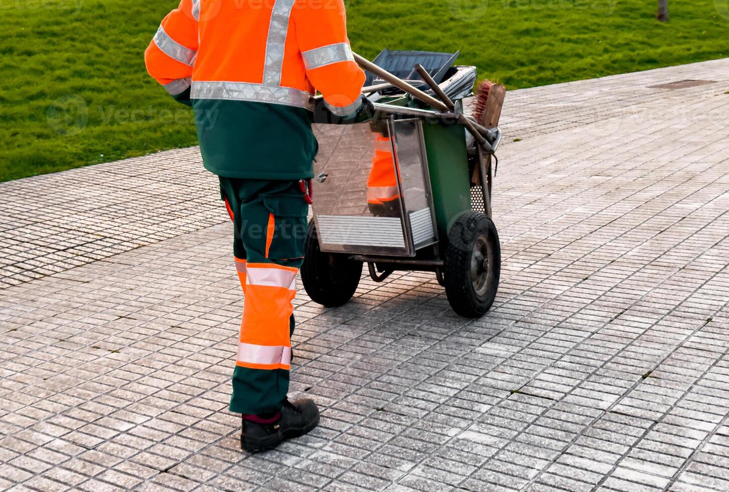 Male sanitation worker pulling cart on park pathway in safety gear photo