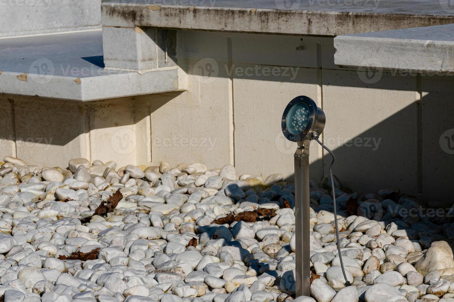 Ground-level garden light illuminates a pebble pathway in daylight photo