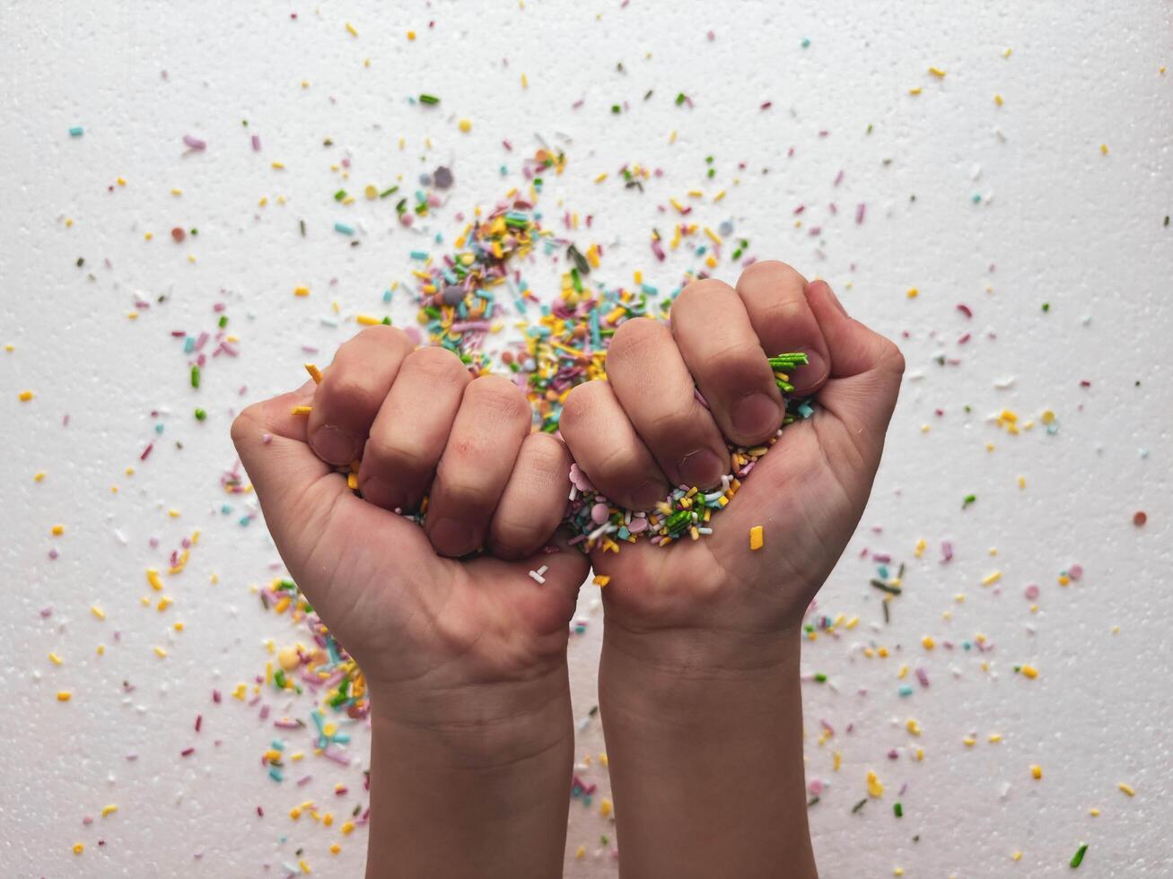 Child's hands clenched into fists with sugar confectionery sprinkles on white background. Child playing at table with small objects for motor skills development. photo