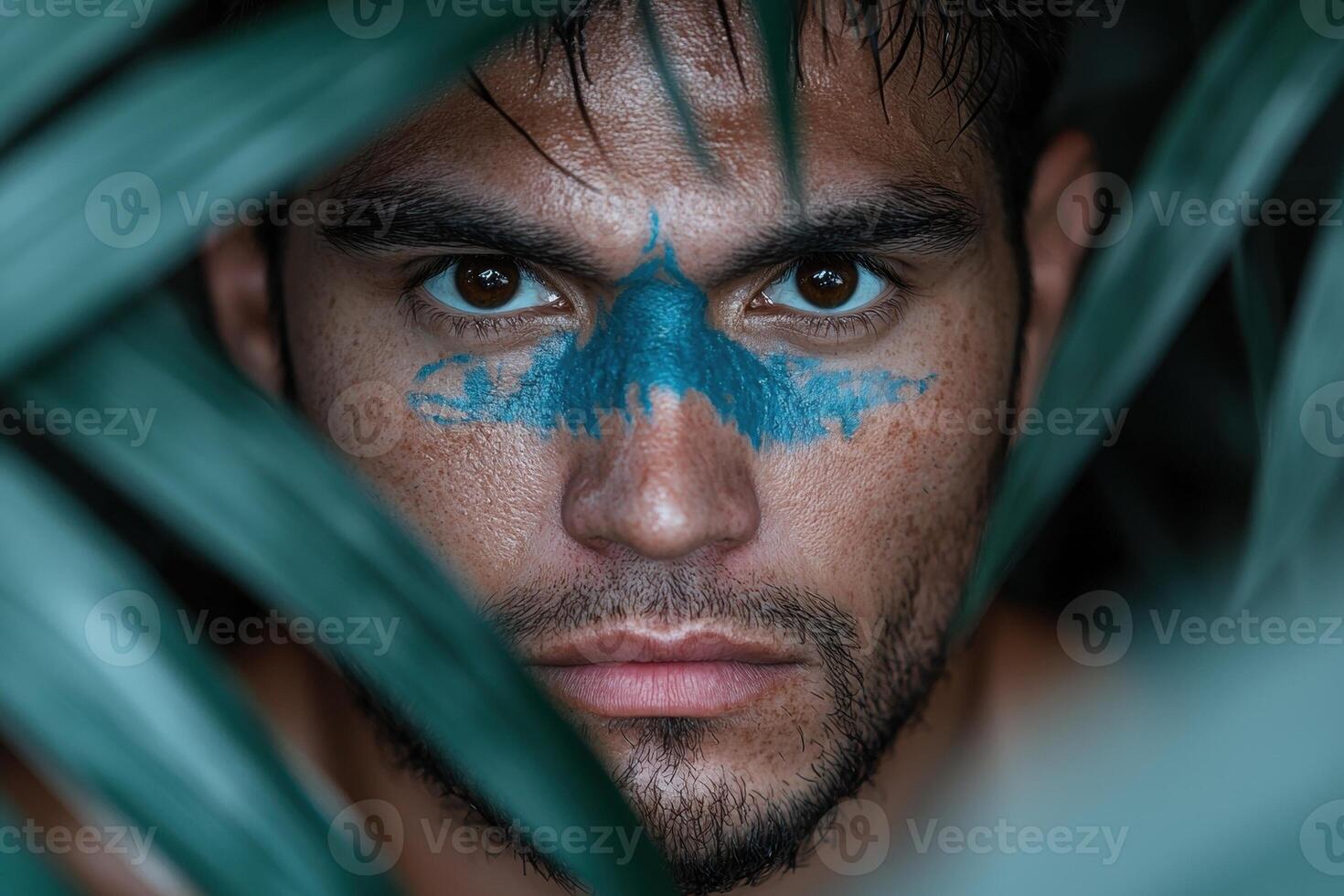 A man with blue face paint peers through green leaves photo