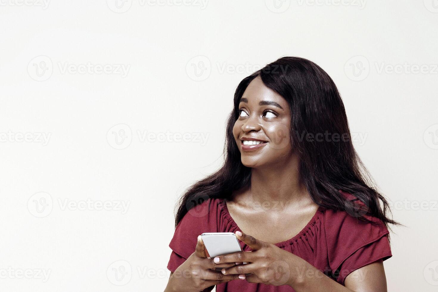 Pretty long-haired young black woman with newest cellphone sitting on floor, looking at copy space over beige studio background, using nice dating, entertaining mobile app on smartphone, panorama photo