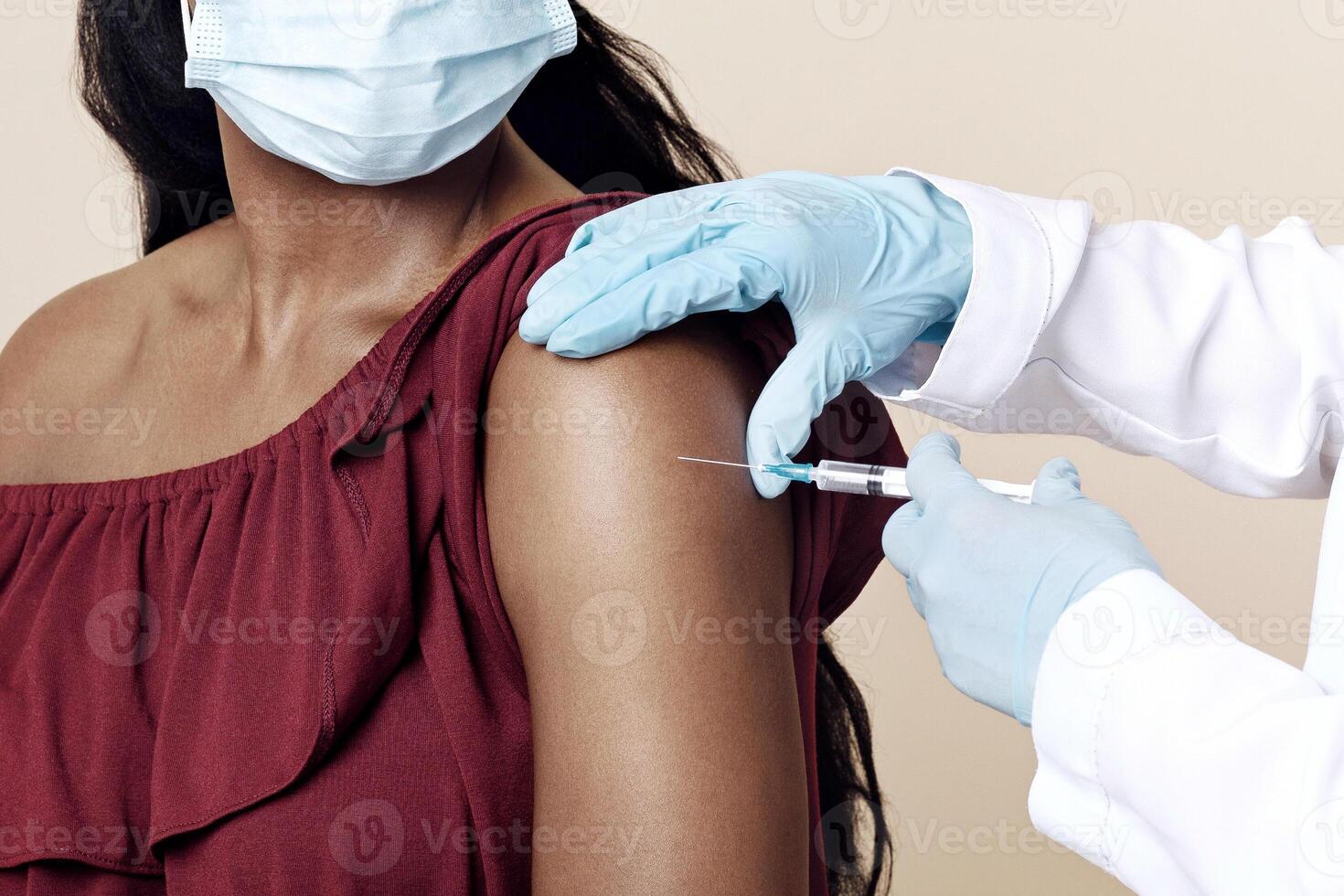 Medical worker wearing gloves and protective face mask making vaccination against coronavirus in shoulder for young black lady, beige studio background, closeup. Safety vaccination concept photo