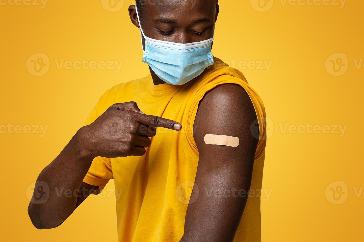 Vaccinated African American Man Wearing Medical Mask Pointing On Arm With Adhesive Bandage After Getting Covid-19 Vaccine Shot, Black Guy Sitting On Chair Over Beige Studio Background, Copy Space photo