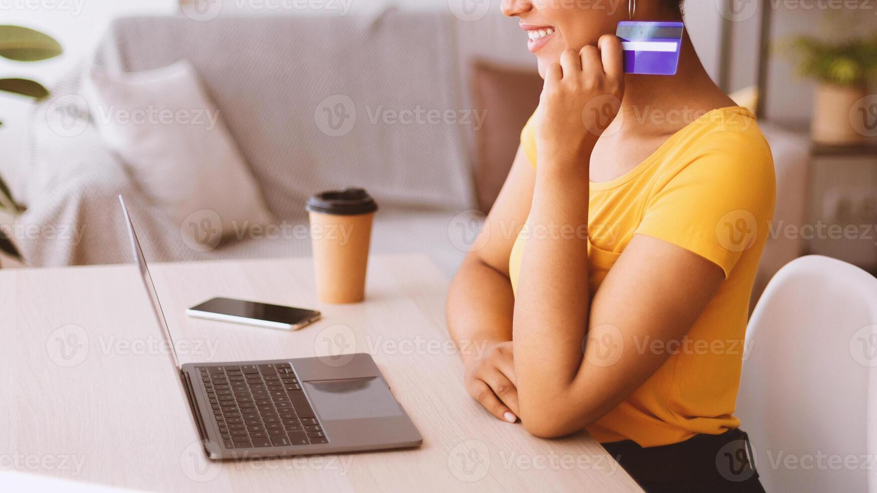 Online Store And Internet Banking Concept. Side view portrait of unrecognizable smiling black lady holding credit card in hand and using laptop, sitting at table with phone and coffee, looking at pc photo